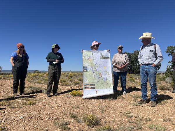 Five ranchers and researchers stand outdoors in a field looking at a large map, discussing land management