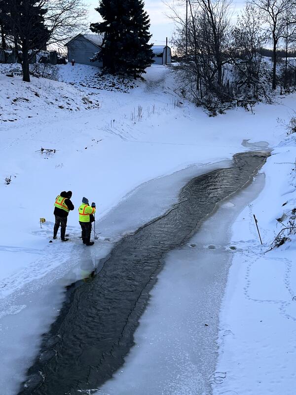Two hydrotechs wearing bright green jackets measure streamflow under frozen river