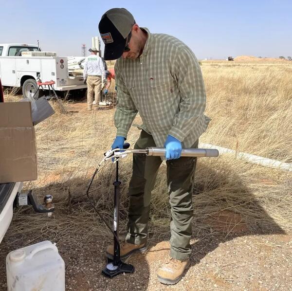 Male in baseball gap, glasses and latex gloved hands standing next to truck in a dry grassy field prepares an instrument
