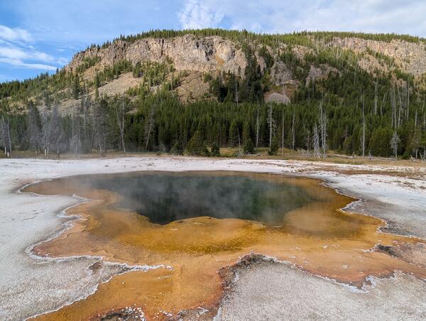 Green hot spring with red edges in a barren white landscape, with trees and a rocky cliff in the background under blue sky
