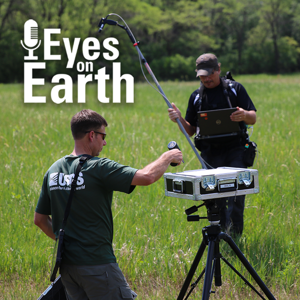 Two men are working with equipment in a grassy area with trees in the background