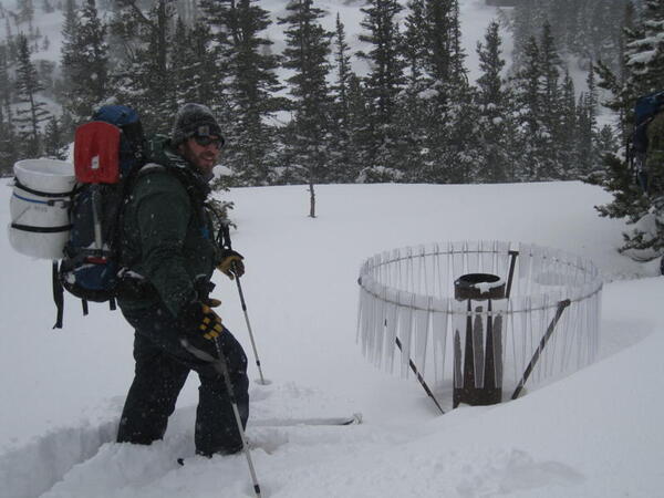 Scientist stands on skis in the snow next to a sample collector.