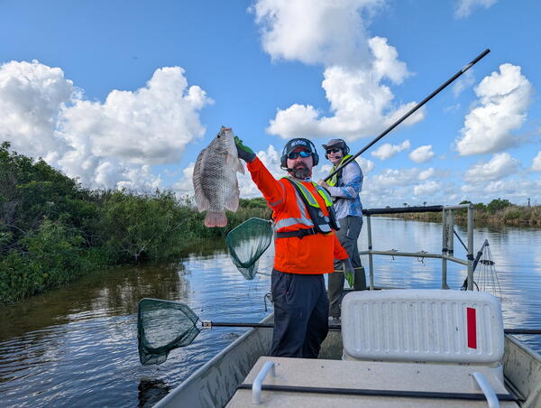 two people in life vests stand in a boat - one holds a fish while the other holds a dipnet