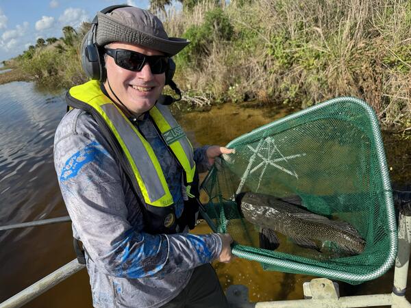 a man holding a fish in a net stands with water and vegetation seen in the background