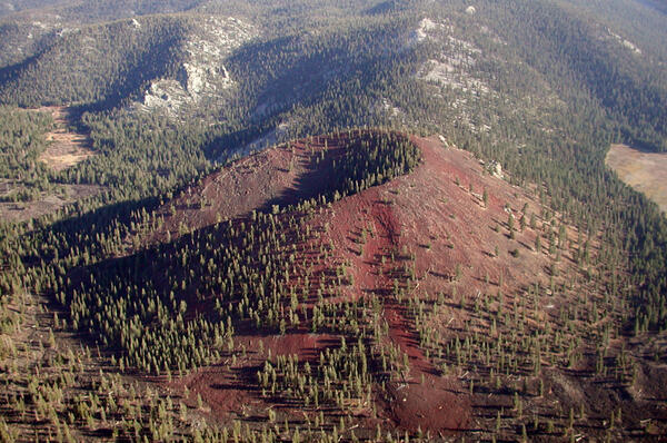 A red scoria cone is dotted with pine forest and sits on the valley floor near low hills of bright white Sierra Nevada granite. 