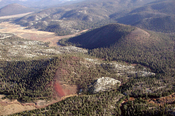 A small forested scoria cone with reddish rocks drapes over bright white outcrops of Sierran granite. The cone sits in a valley amid low rolling hills backed by higher granite peaks. 