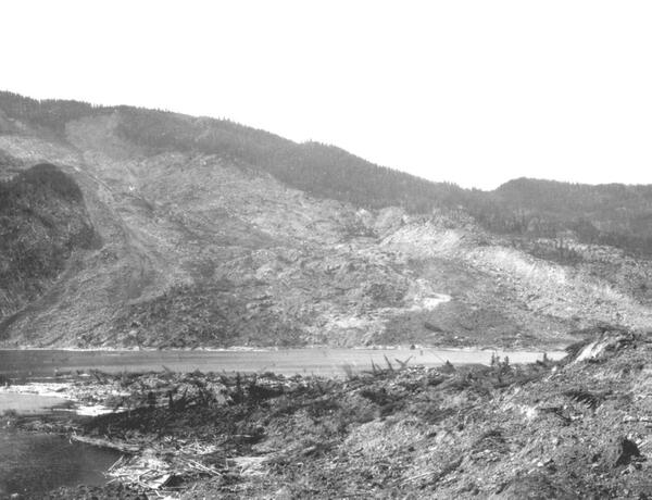 Black and white photo of a hillside that has been devastated by a landslide, with rock debris in the foreground and a lake