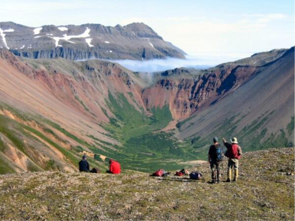 Two people standing by a ledge facing and gesturing away towards mountains in the distance; packs and two people on ground