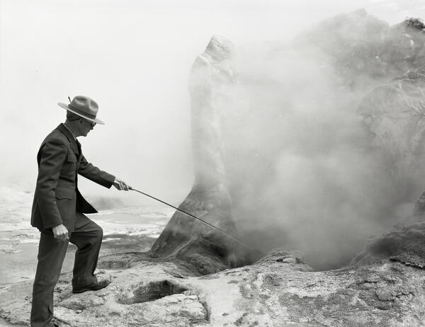 Black and white photo of a park ranger using a probe to take a temperature from a geyser cone, which is obscured by steam