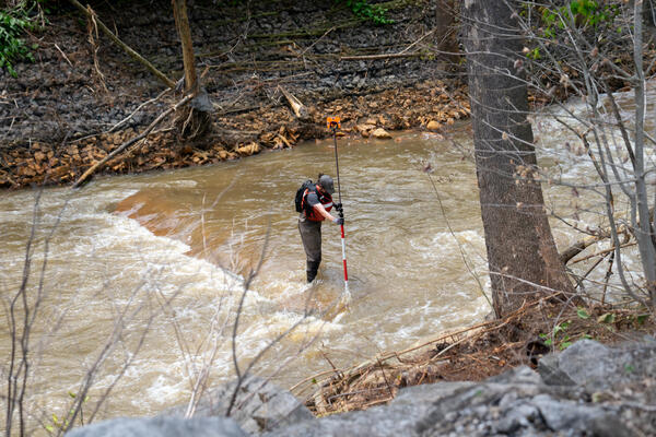 A man stands in a stream with surveying equipment.