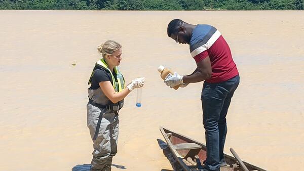 USGS scientist standing in shallow brown river training Ghanaian scientist standing on wood canoe on water sample collection