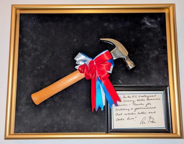 Hammer with a red, white, and blue bow mounted on black felt canvas with a golden frame