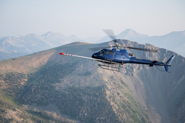 A blue helicopter flies in front of mountain with a long, white boom arm sticking out the front of the aircraft with red tip