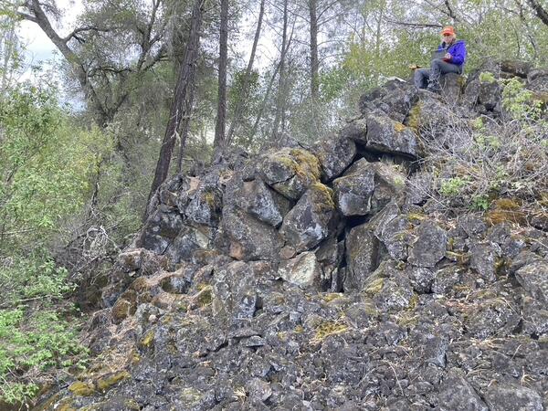 A female geologist perches on a mottled gray and white boulder at the top of a pile of the same type of rock