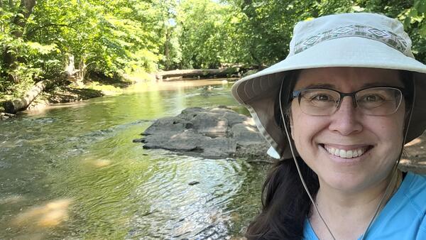 A woman in a tan hat is smiling for her picture while standing beside a river in the forest.