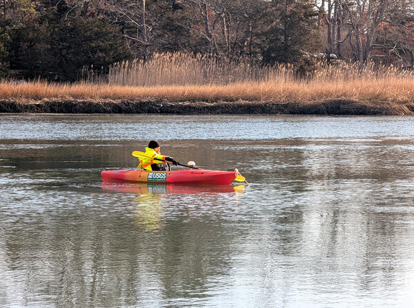 A man in a neon yellow jacket on an orange kayak in a river holding a paddle.