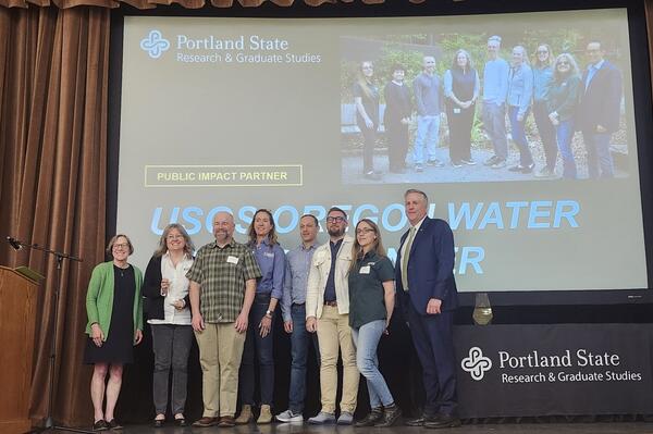 8 people stand on a stage in front of a projection screen with award info on it