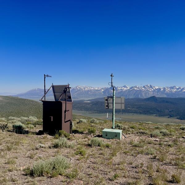 In this photo, a large brown metal box and a small aqua-colored box are both topped by antenna poles and solar panels. They sit side-by-side on a sagebrush-covered hill overlooking a broad valley and a line of jagged, snow-capped mountains.