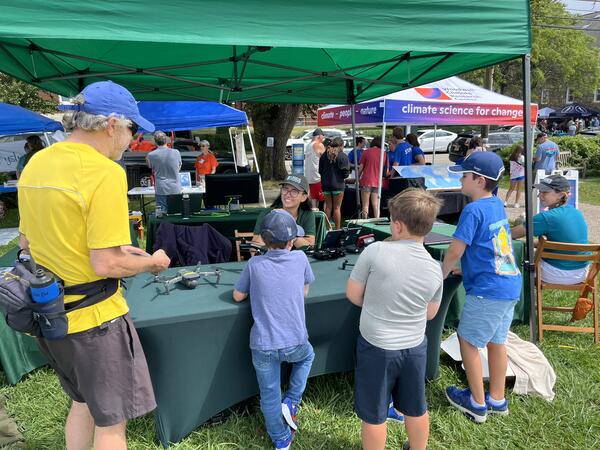 USGS scientist sitting outside behind a table with drones on it, three children and an adult standing in front talking to her