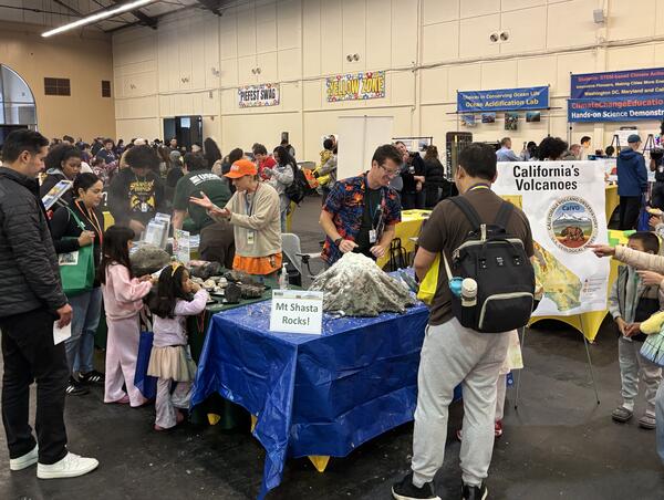 This photo shows a science festival booth surrounded by children and their families. A model volcano sits on a blue tarp behind a "Mt Shasta Rocks!" sign. Behind the table, a scientist in an orange USGS cap explains volcanic rocks, while another scientist in a volcano-themed Hawaiian shirt prepares the model volcano for its next "eruption".