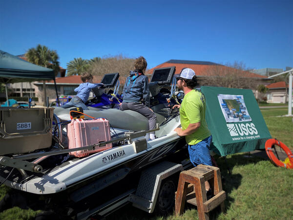 Two children sit on personal watercraft over land in a park while USGS staff explain coastal mapping