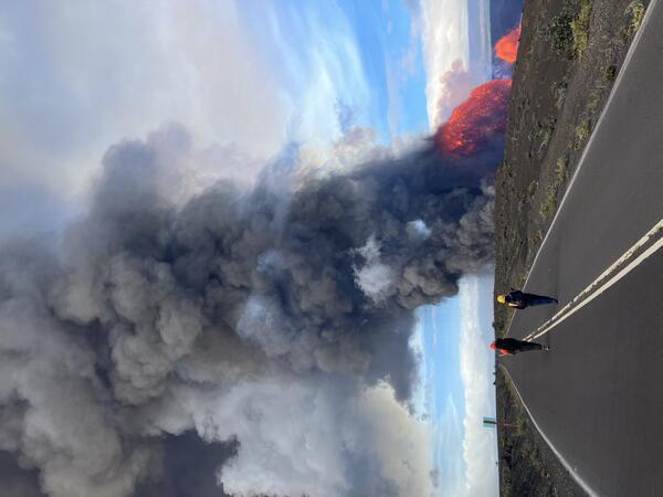 Color photograph of scientists walking on a closed road with volcanic eruption in the distance