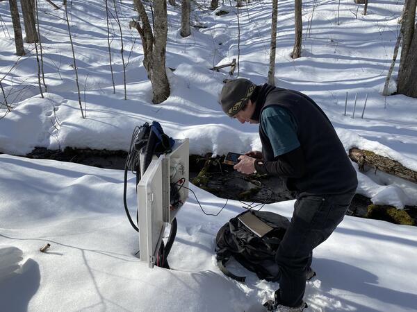 A man in the woods in the snow looks at a data logger.