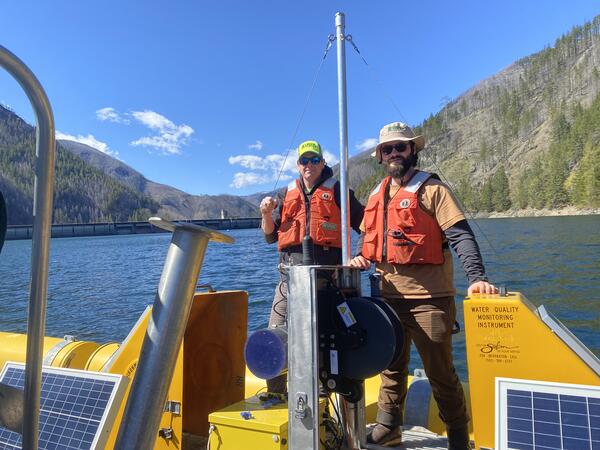 two males stand in safety gear and hats stand on a bright yellow floating platform in blue lake water on a sunny day