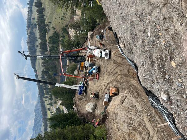 Several pieces of large construction equipment, including two cranes and a cement mixer, in a cleared area among trees.  A grass and tree-covered hill rises in the background.