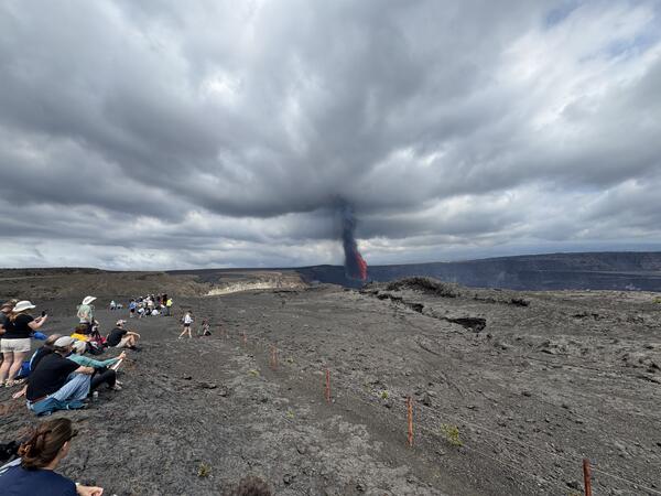Color photograph of people sitting on the ground with an eruption visible in the background