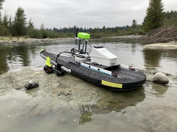 A small USV sits on a shallow riverbank, equipped with sensors, a propulsion unit, and a compact central hull