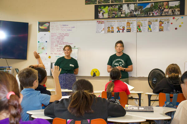 Two people stand at the front of a classroom of children. 