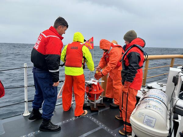 Four people on ship in ocean, huddled around a piece of equipment near railing