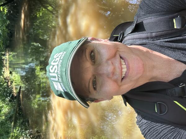 A smiling woman wearing a green USGS baseball hat standing in water and wearing a camera backpack. 