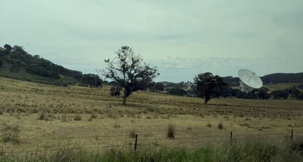 A large antenna reaching up to a cloudy sky with a few trees and buildings in the background