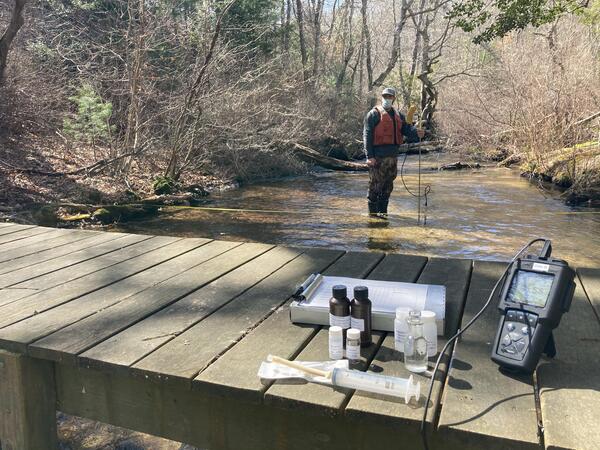 A man stands in a shallow river with a flow meter in the background and water quality equipment in the foreground on a dock.