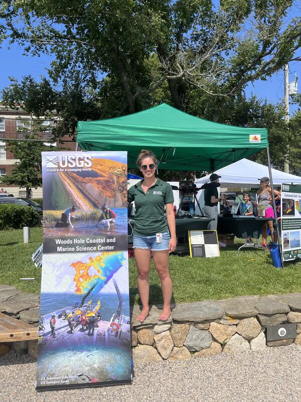 Person standing next to a vertical banner outside on a nice day at an event with people and a tent in the background