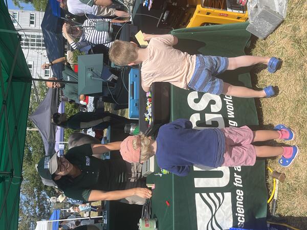 Person in USGS hat and shirt at USGS booth outside smiling at kids playing with our Legos