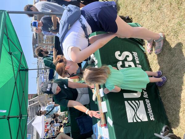 woman showing adult and child a replica sediment core on table outside under green tent