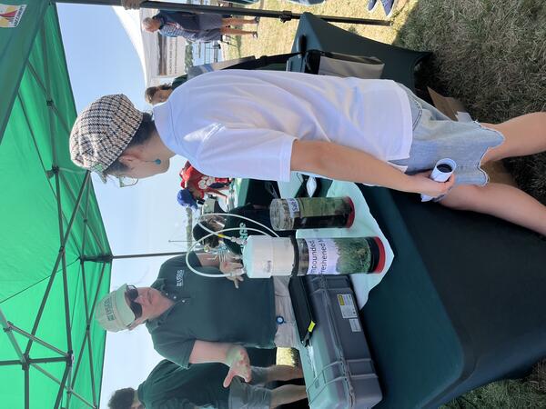 Person in USGS shirt at booth under tent outside talking to person while referencing equipment on the table