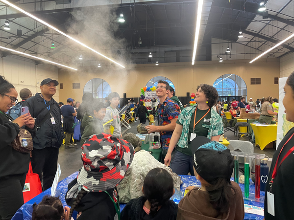 This photograph shows a model volcano sitting on a table surrounded by people. The model volcano has a plume of fine material suspended in the air above it, indicating it erupted immediately before the photo was taken. In front of the table, children participants face away from the camera looking at the volcano and the plume. Behind the table, animated scientists in brightly colored shirts are smiling and looking up at the plume.