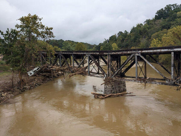 A photo showing a bridged spanning a muddy river. The bridge is covered with logs and dirt caused by recent flooding. 