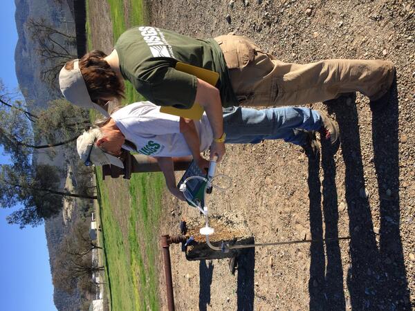 Two USGS scientists  wearing USGS shirts and baseball caps stand next to a rusting wellhead pipe and concrete well casing on a gravel field. One holds a gas analyzer with tubing; the other takes notes on a yellow clipboard. A green valley and low mountain ridges rise in the background under a clear blue sky.