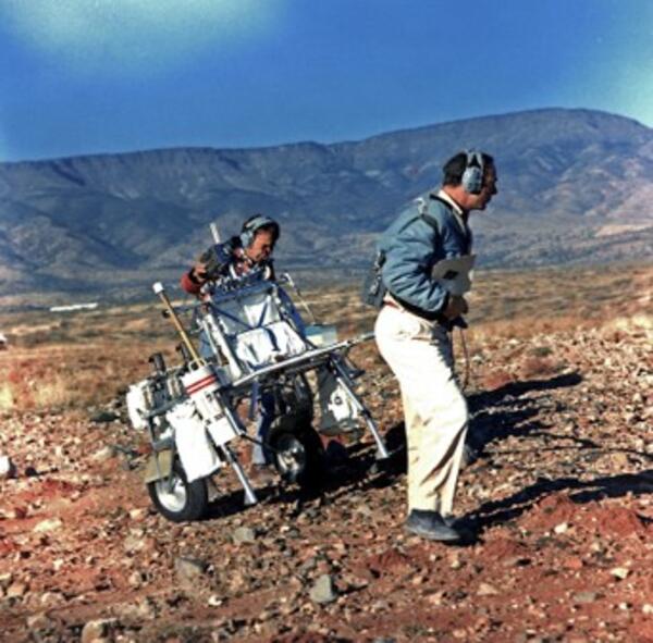 Two men wearing jackets and headsets pulling a piece of equpment up a moderate slope in the desert southwest