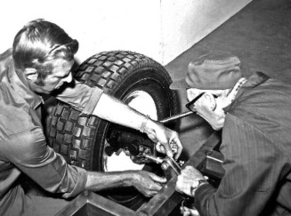A black and white photo of two men working on the suspension of an open chassis vehicle