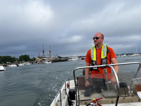 Man with life preserver driving boat with large ship in the background.