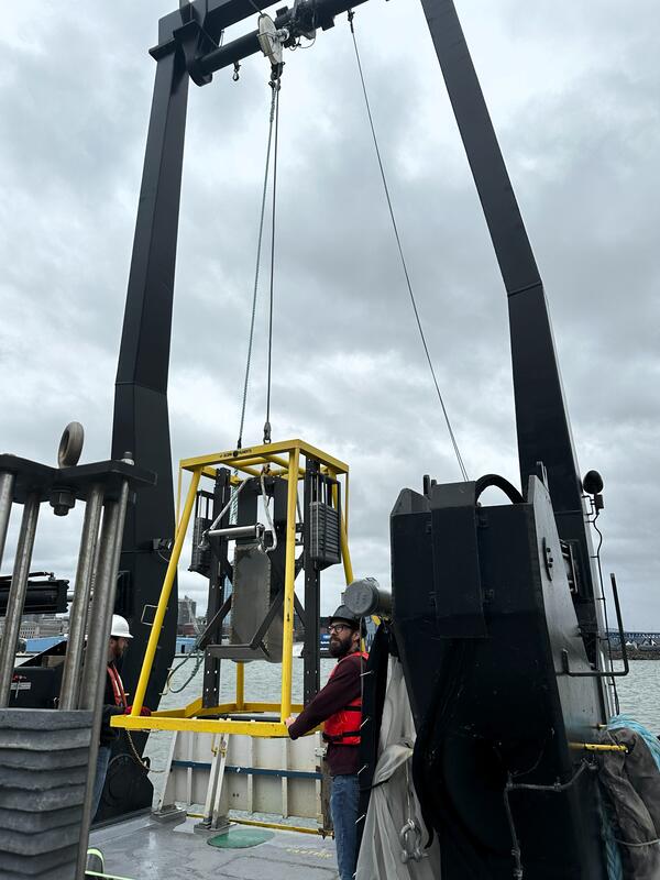 Two scientists guide sampling equipment attached to large crane, on to deck of research vessel