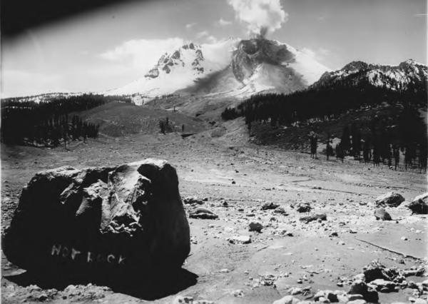 A black and white photograph looks across a swath cut through a pine forest by a debris and mud flow that originates at a steaming volcanic peak in the distance. A large crater is visible on the side of the volcano, which is emitting the steam. The rest of the volcano is heavily blanketed in snow.