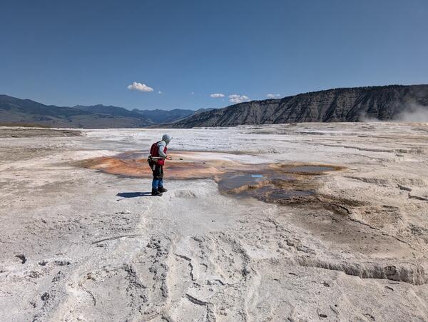 Woman in red vest standing on white terrace in front of reddish hot spring under blue sky. Mountains in the background.