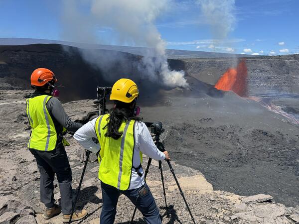 color photo of two geologist looking at the lava fountain in the background.